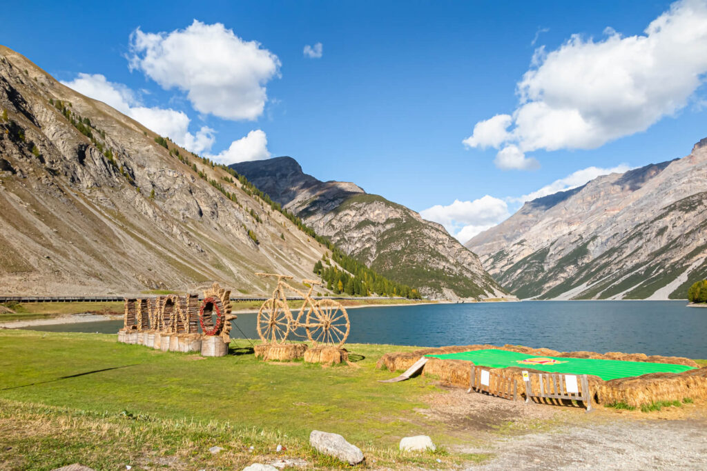 A scenic lake surrounded by mountains with a straw sculpture and a green field in the foreground.