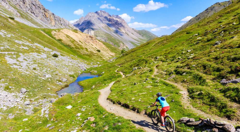 A cyclist rides a mountain trail with alpine scenery.